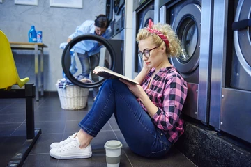 Young woman sitting inside a laundromat and reading a book